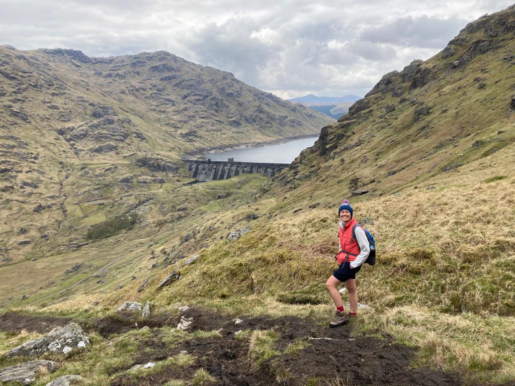 Photo of Jemima standing in the hills in front of a dam/loch. She is wearing shorts, a woolly hat, an orange body warmer and hiking boots.