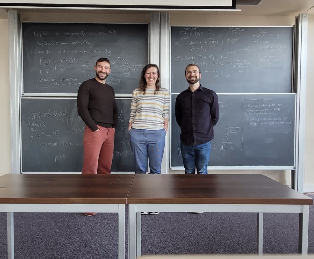 Photo of 3 people in front of a blackboard. Jemima is standing in the middle and smiling